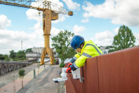 A worker in a bright yellow jacket and blue helmet operates equipment attached to a brown railing outdoors, with a large yellow crane and cloudy sky in the background.
