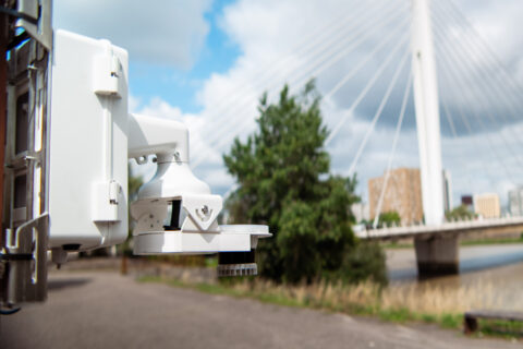 A close-up of a white security camera mounted outdoors, with a blurred background showing a cable-stayed bridge, trees, and buildings under a partly cloudy sky.
