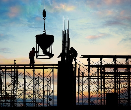 Silhouetted construction workers on scaffolding operate machinery and work on a building framework at sunset, with a crane lifting a concrete bucket against a colorful sky.