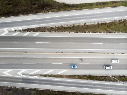 Aerial view of a divided highway with three cars traveling in opposite directions. The roads are separated by a median with sparse vegetation. Road markings and arrows are visible on the asphalt.
