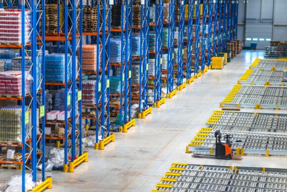 A large warehouse with tall blue racks filled with stacked goods and pallets. An orange and black forklift is parked in an open area next to roller conveyor lanes, ready for loading or unloading items.