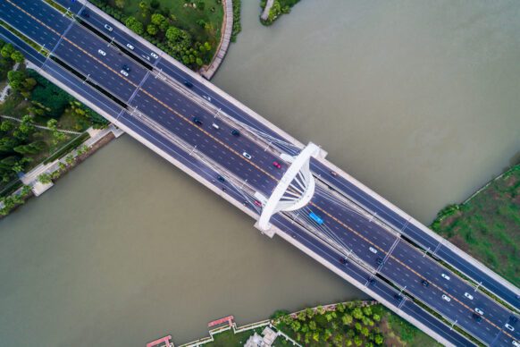 Aerial view of a wide, modern bridge with multiple lanes for vehicles, crossing over a river. Cars and trucks are visible on the bridge, and green trees line the riverbanks on both sides.