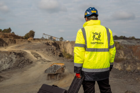Man wearing a yellow safety outfit and an Inspemon jacket, standing in front of a construction site.