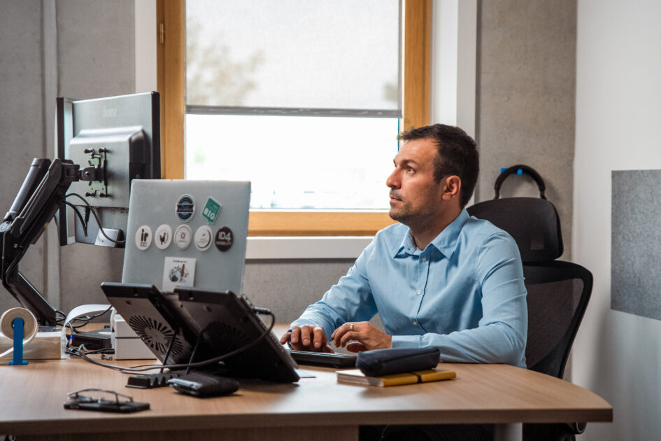 Man in a blue shirt works at a desk with dual monitors and a laptop. He looks focused, sitting in a modern office with a window and gray walls.