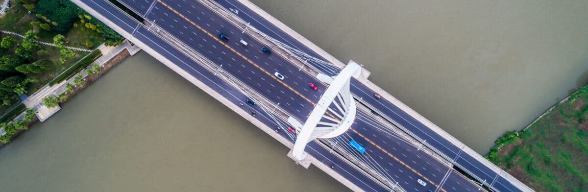Aerial view of a wide bridge with multiple lanes for vehicles crossing over a river, surrounded by greenery on both sides.