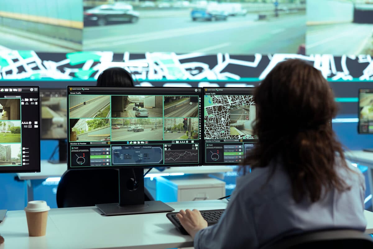 Two people sit at desks in a control room, monitoring multiple surveillance camera feeds and maps displayed on large computer screens, with a coffee cup beside one of the monitors.